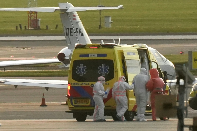 Une personne en combinaison de protection (2e d) escortée vers une ambulance depuis un avion médicalisé transportant des passagers du bateau de croisière MV Hondius, soupçonnés d'être infectés par le hantavirus, à l'aéroport de Schiphol, le 6 mai 2026 près d'Amsterdam, aux Pays-Bas
