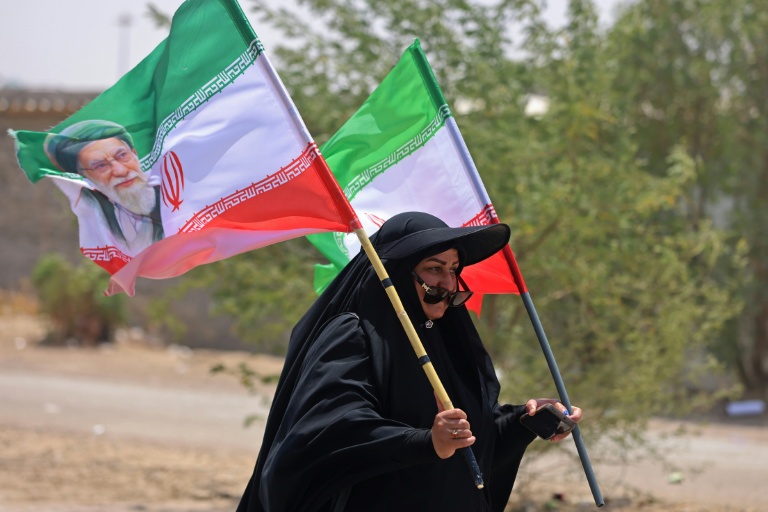 Une pèlerine iranienne brandit des drapeaux iraniens dont l'un comprend un portrait du guide suprême Ali Khamenei, tué dans un bombardement américain, le 29 juillet 2026 dans la ville-frontière de Zurbatiyah, en Irak