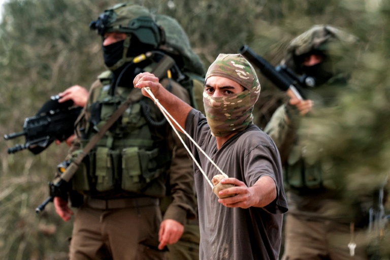 Des soldats israéliens derrière un homme masqué lançant des pierres sur des Palestiniens rassemblés pour la saison annuelle de la récolte des olives, lors d'une attaque de colons israéliens sur le village palestinien de Beita, au sud de Naplouse, en Cisjordanie occupée, le 10 octobre 2025